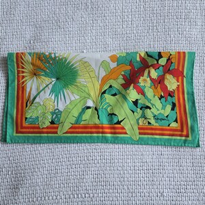 May include: Rectangular silk scarf with a tropical floral and foliage design. The scarf features large green leaves, red and yellow flowers, and a green border with orange and red stripes. The text "Jim Thompson" is visible.
