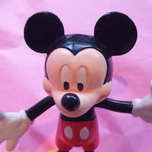 Peut inclure: Figurine de Mickey Mouse avec des oreilles noires, un visage noir et un museau beige. La figurine porte un short rouge avec deux boutons blancs, des chaussures jaunes et des gants blancs. Le fond est rose clair.