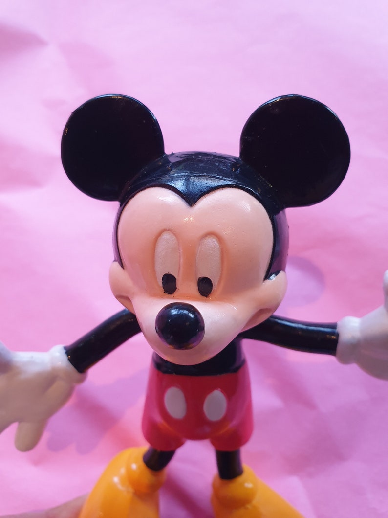 Peut inclure: Figurine de Mickey Mouse avec des oreilles noires, un visage noir et un museau beige. La figurine porte un short rouge avec deux boutons blancs, des chaussures jaunes et des gants blancs. Le fond est rose clair.