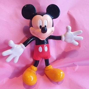 Peut inclure: Figurine de Mickey Mouse avec des oreilles noires, une t&ecirc;te noire et un visage blanc. La figurine porte un short rouge, des chaussures jaunes et des gants blancs. La figurine est pos&eacute;e avec les bras tendus sur un fond rose.