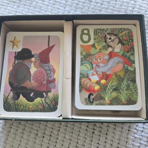 May include: A deck of playing cards inside a dark green box. The left card shows two gnomes on a swing with a gold star. The right card features the number 8, a gnome with a wheelbarrow of food, and a dog in a garden setting with green plants.