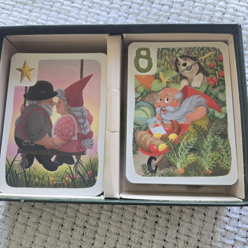 May include: A deck of playing cards inside a dark green box. The left card shows two gnomes on a swing with a gold star. The right card features the number 8, a gnome with a wheelbarrow of food, and a dog in a garden setting with green plants.