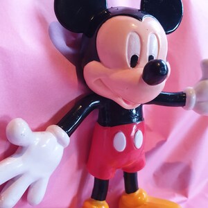 Peut inclure: Figurine de Mickey Mouse avec une t&ecirc;te et des oreilles noires, un visage rose et un corps noir. La figurine porte un short rouge avec des boutons blancs et des chaussures jaunes. Le fond est rose.