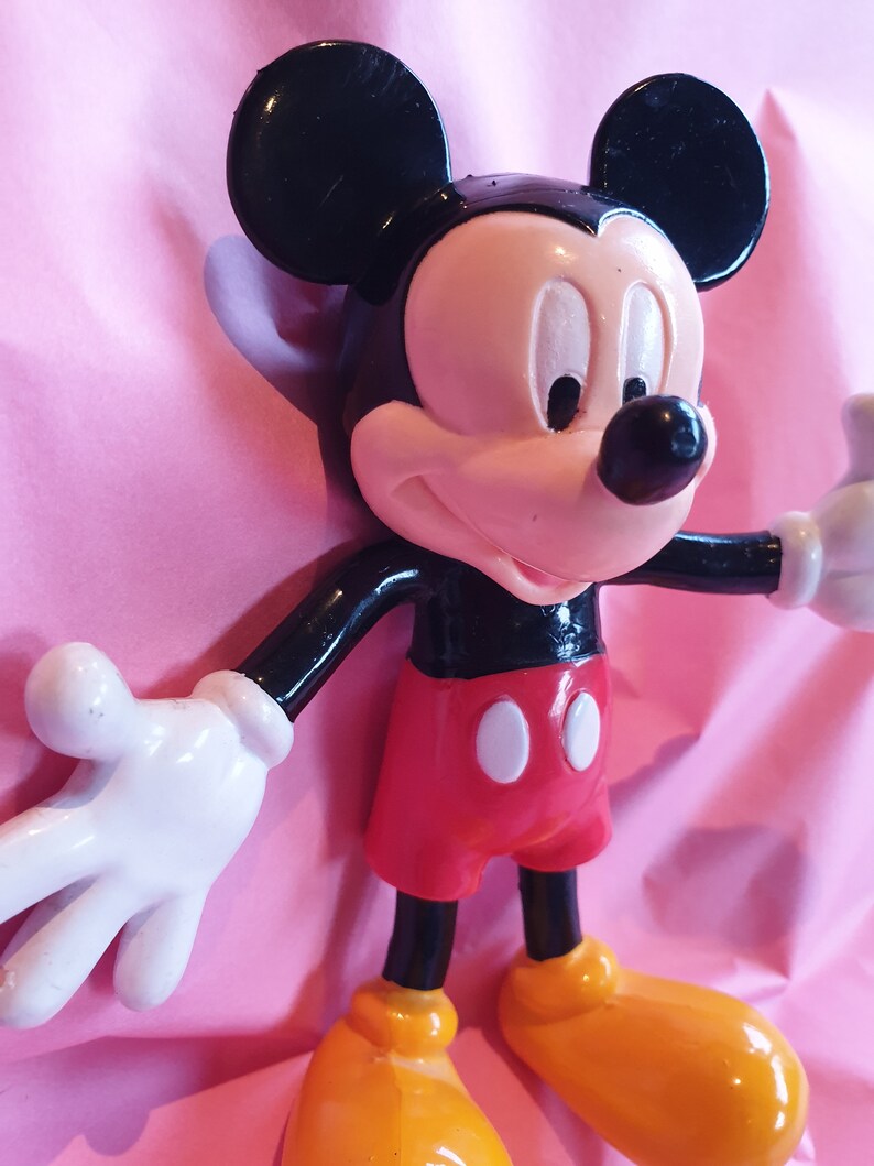 Peut inclure: Figurine de Mickey Mouse avec une t&ecirc;te et des oreilles noires, un visage rose et un corps noir. La figurine porte un short rouge avec des boutons blancs et des chaussures jaunes. Le fond est rose.