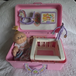 Può includere: Un set di gioco vintage per bambole Cabbage Patch Kids rosa. Il set include una bambola, una culla, un cavallo a dondolo e un piccolo giocattolo. La valigia è rosa con una maniglia e dettagli decorativi. La bambola ha i capelli biondi e indossa un vestito.