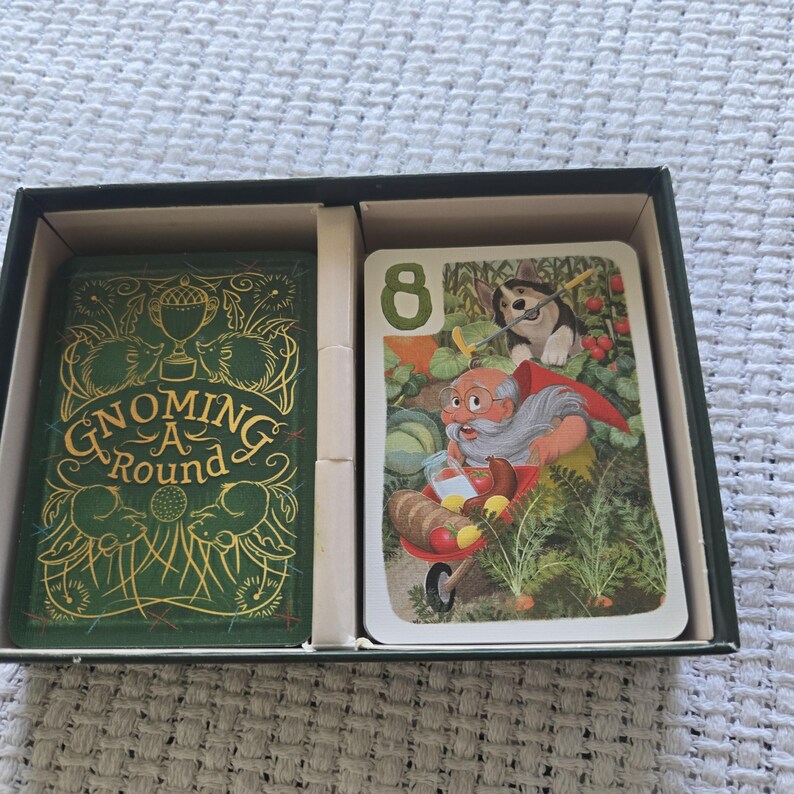 May include: A Gnoming A Round card game set in a black box. The left side features a green card with gold illustrations of hedgehogs and a trophy. The right side shows a card with the number 8, a gnome with a white beard, and a dog in a garden.