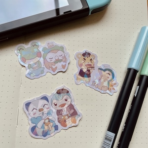 May include: Four holographic stickers featuring animal characters from the video game Animal Crossing. The stickers feature a frog holding a cup of coffee, an owl holding a slice of cake, a bear wearing a beret and eating a slice of cake, and a penguin wearing a beret and holding a cup of coffee. The stickers are on a white background with a dotted pattern.