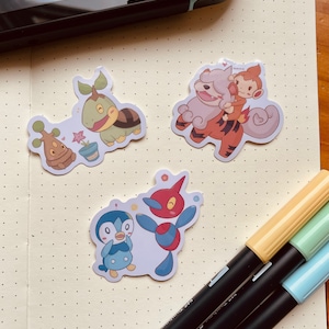 May include: Four colorful cartoon character stickers featuring Pokemon characters. The stickers are on a white dotted notebook page. The characters are a brown and green turtle, a brown and white dog, a red and blue bird, and a blue and white penguin.