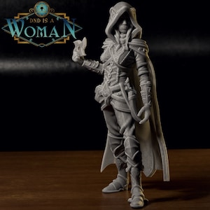 May include: A gray 3D printed miniature of a female figure wearing a hooded cloak and holding a dagger. The figure is standing on a wooden surface.