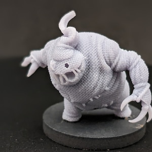 May include: A light gray, 3D-printed monster figurine with a textured, burlap-like appearance. The creature has clawed hands, a toothy mouth, and a spiraling head tendril. It stands on a dark gray circular base.