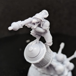May include: A white plastic miniature figurine of a creature with a helmet and a large gun. The creature is standing on a barrel.