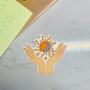 May include: A sticker with a sun and moon design, surrounded by stars, held in two hands. The sun is orange and yellow, the moon is blue and white, and the stars are yellow.