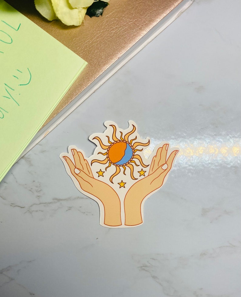 May include: A sticker with a sun and moon design, surrounded by stars, held in two hands. The sun is orange and yellow, the moon is blue and white, and the stars are yellow.