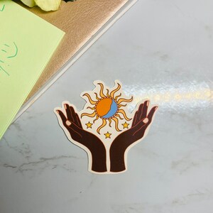 May include: A sticker with a pair of brown hands holding a sun and moon design. The sun is orange and yellow with a blue crescent moon inside. There are five small yellow stars around the sun and moon.