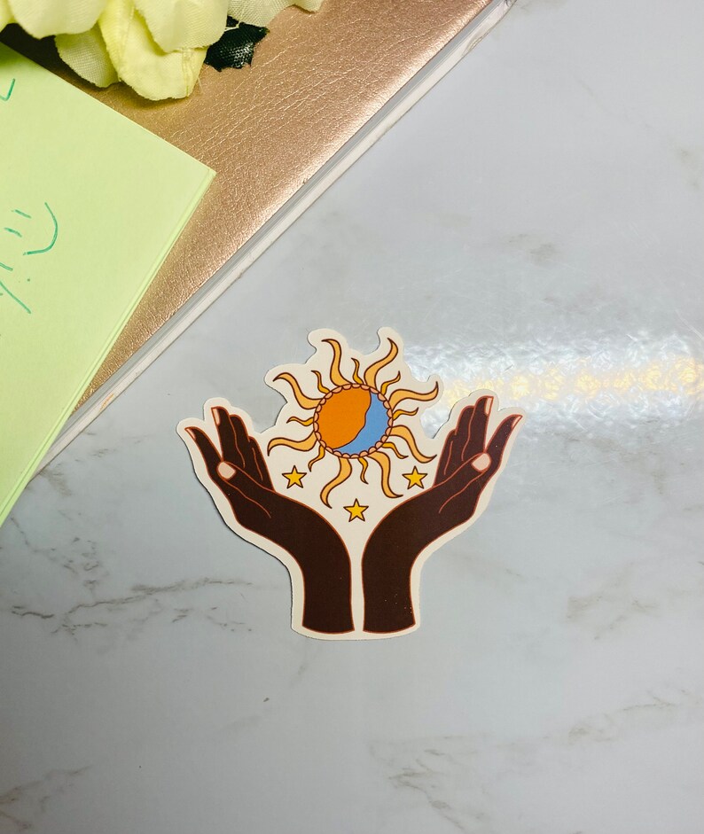 May include: A sticker with a pair of brown hands holding a sun and moon design. The sun is orange and yellow with a blue crescent moon inside. There are five small yellow stars around the sun and moon.