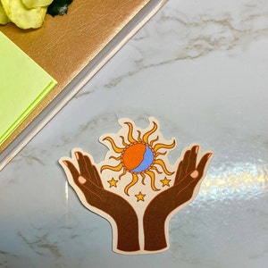 May include: A sticker with two brown hands holding a sun and moon design. The sun is orange and yellow with a blue crescent moon inside. There are three small yellow stars around the sun and moon.