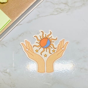 May include: A sticker with a sun and moon design, surrounded by stars, held in two hands. The sun is orange and yellow, the moon is blue and white, and the stars are white.