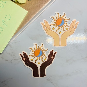May include: Two sticker designs featuring a sun and moon with stars, each held by two hands. One design has light skin tone hands, the other has dark skin tone hands.