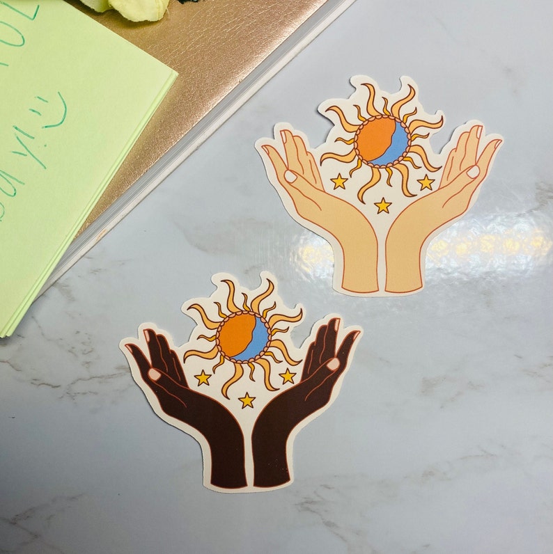 May include: Two sticker designs featuring a sun and moon with stars, each held by two hands. One design has light skin tone hands, the other has dark skin tone hands.