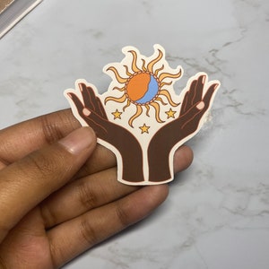 May include: A sticker with a stylized illustration of two hands holding a sun with a crescent moon inside. The sun is orange and yellow, the moon is blue, and the hands are brown. There are five white stars around the sun and moon.