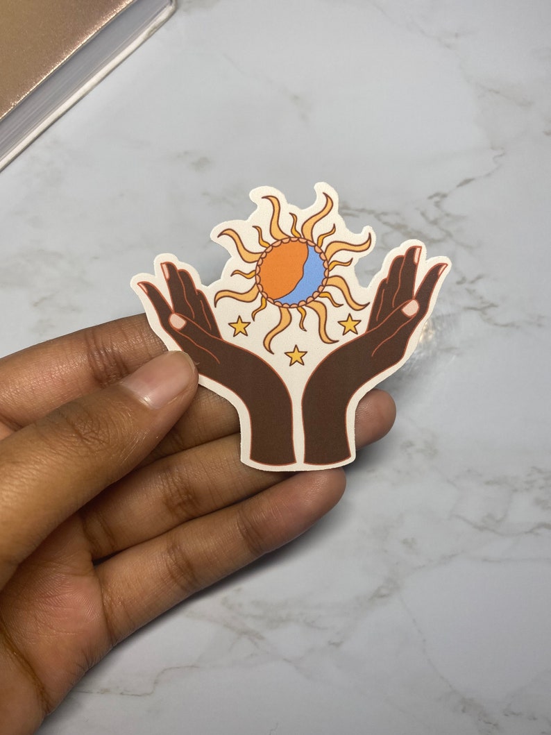 May include: A sticker with a stylized illustration of two hands holding a sun with a crescent moon inside. The sun is orange and yellow, the moon is blue, and the hands are brown. There are five white stars around the sun and moon.