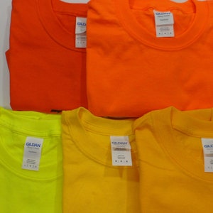 May include: Four Gildan brand t-shirts in different colors: orange, yellow, neon yellow, and a darker orange. The shirts are folded and stacked on a white surface.