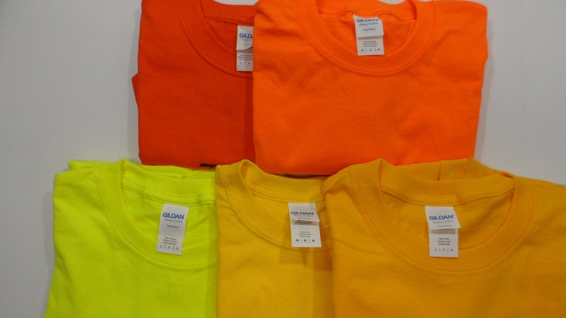 May include: Four Gildan brand t-shirts in different colors: orange, yellow, neon yellow, and a darker orange. The shirts are folded and stacked on a white surface.