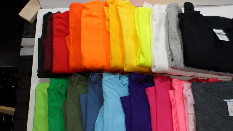 May include: A variety of plain t-shirts in different colors, folded and stacked on a white surface.