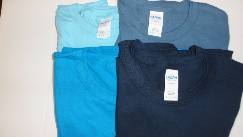 May include: Four folded Gildan brand t-shirts in different shades of blue: light blue, turquoise, denim blue, and navy blue.