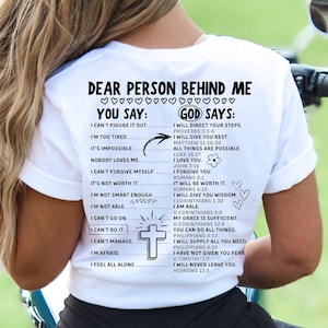 May include: White t-shirt with a black graphic print of a cross and text that reads "Dear person behind me. You say: I can't figure it out. I'm too tired. It's impossible. Nobody loves me. I can't forgive myself. It's not worth it. I'm not smart enough. I'm not able. I can't go on. I can't do it. I can't manage. I'm afraid. I feel all alone. God says: I will direct your steps. I will give you rest. All things are possible. I love you. I forgive you. It will be worth it. I will give you wisdom. I am able. My grace is sufficient. You can do all things. I will supply all you need. I have not given you fear. I will never leave you."
