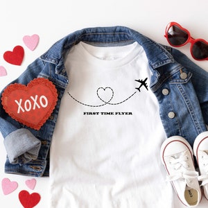 May include: White baby t-shirt with a black airplane and heart design. The text "First Time Flyer" is printed below the design. The t-shirt is laid out on a white surface with a denim jacket, red heart-shaped sunglasses, and red and pink felt hearts.