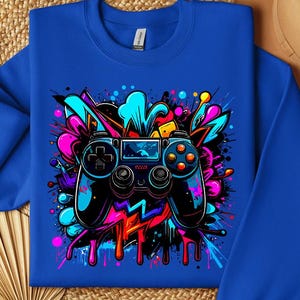 May include: Royal blue sweatshirt featuring a vibrant graphic of a video game controller. The controller is black and blue, surrounded by colorful paint splatters in pink, teal, and yellow. The design has a graffiti art style.