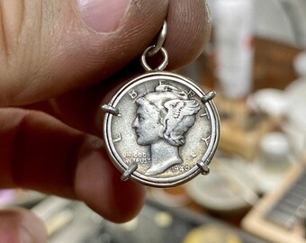 Może przedstawiać: Srebrzysty wisiorek z monetą Mercury dime w metalowej oprawie z czterema ząbkami. Moneta przedstawia profil Liberty w skrzydlatej czapce, z napisem LIBERTY i IN GOD WE TRUST oraz rokiem 1940 widocznym w dolnej części monety.
