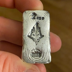 Può includere: Un lingotto d'argento rettangolare con una superficie testurizzata che ricorda le venature del legno. La parte superiore presenta il numero 2, un emblema stilizzato di testa di lupo e la scritta 999FS impressa nel metallo con bordi arrotondati.