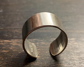 Może przedstawiać: Szeroki, otwarty pierścionek z metalu w kolorze srebrnym o gładkim, szczotkowanym wykończeniu. Obrączka ma prosty, pozbawiony zdobień design z przerwą z tyłu, co umożliwia regulację rozmiaru. Widoczna jest wewnętrzna powierzchnia metalowej obrączki.