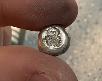 Może przedstawiać: Mały, okrągły, srebrny metalowy żeton z wytłoczonym wzorem czaszki i skrzyżowanych piszczeli. Powierzchnia metalu ma lekko nieregularny, ręcznie odlewany wygląd z polerowanym wykończeniem na wypukłych detalach.