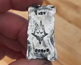 May include: A rectangular silver bar with a rough, hand-poured texture. The front features a stamped wolf head logo, the text 1 oz, and the text 999FS. The metal has a metallic sheen with dark oxidation in the recessed areas of the design.