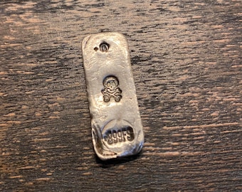 May include: A rectangular silver bar with a textured, metallic finish. The bar features a stamped skull and crossbones icon in the center, a small circular mark with the number 5 at the top, and the text 999FS stamped at the bottom.