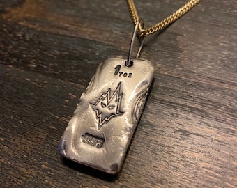 May include: A rectangular silver pendant on a gold-coloured chain. The pendant features a stamped graphic of a stylised face with sharp features, the text 1 TOZ, and the text 999FS. The metal surface has a textured, hand-poured appearance.