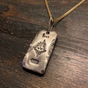 Può includere: Un ciondolo rettangolare in argento su una catena color oro. Il ciondolo presenta un grafico stampato di un volto stilizzato con tratti affilati, il testo 1 TOZ e il testo 999FS. La superficie metallica ha un aspetto testurizzato.