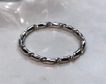 Może przedstawiać: Cienki, polerowany pierścionek w kolorze srebrnym z ciągłą teksturą skręconego sznura lub spirali wokół całej obrączki. Metalowa powierzchnia jest odblaskowa i gładka, tworząc powtarzający się wzór ukośnych segmentów wzdłuż pierścionka.