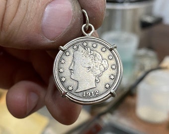 Może przedstawiać: Srebrzysty wisiorek w kształcie monety z profilem Liberty w koronie z napisem LIBERTY, otoczony trzynastoma gwiazdami i rokiem 1912 u dołu, umieszczony w okrągłej metalowej oprawie z uchwytem do zawieszenia na górze.