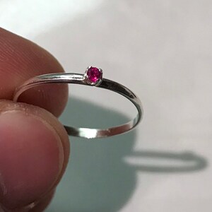 May include: A thin, polished silver-toned metal ring featuring a single, small, round-cut vibrant pink gemstone set in a simple four-prong setting. The band is smooth and slender, showcasing a minimalist design with a bright, reflective finish.
