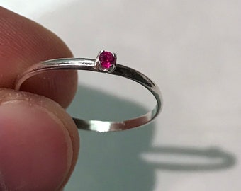 May include: A thin, polished silver-toned metal ring featuring a single, small, round-cut vibrant pink gemstone set in a simple four-prong setting. The band is smooth and slender, showcasing a minimalist design with a bright, reflective finish.