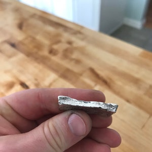 Puede incluir: Una peque&ntilde;a pieza de metal de color plateado con forma irregular, una superficie rugosa y texturizada, y bordes desiguales y dentados. El objeto tiene un brillo met&aacute;lico y parece ser un fragmento s&oacute;lido fundido con una parte superior plana.