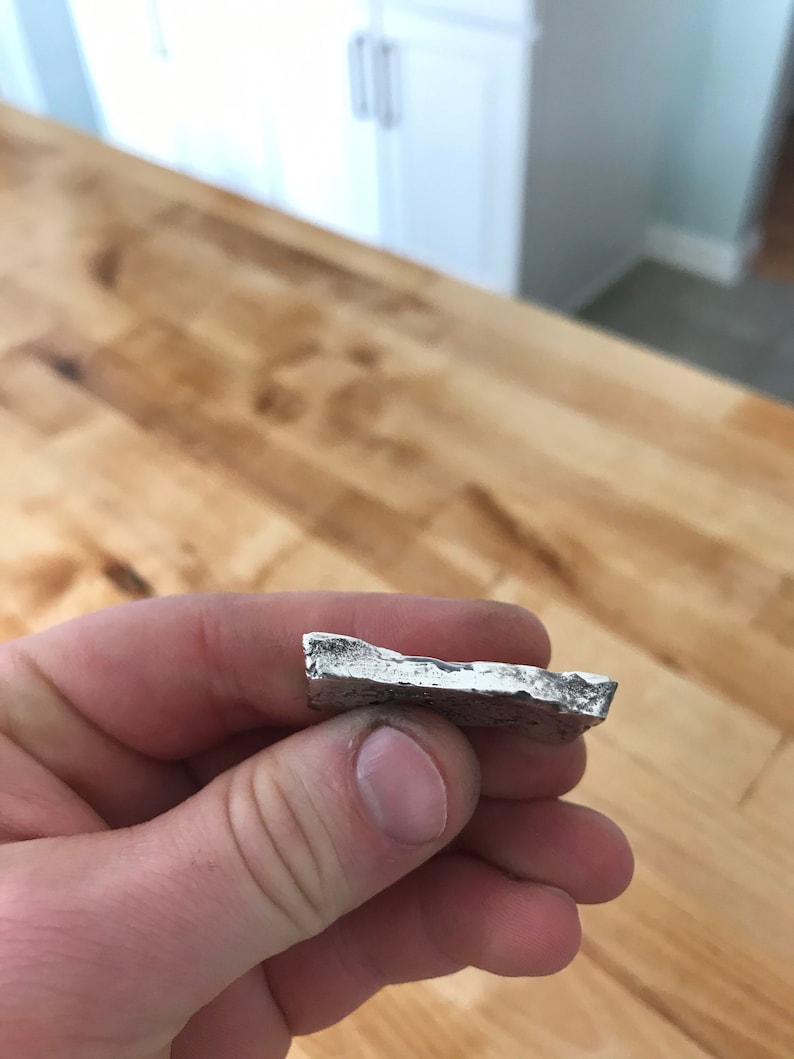 Puede incluir: Una peque&ntilde;a pieza de metal de color plateado con forma irregular, una superficie rugosa y texturizada, y bordes desiguales y dentados. El objeto tiene un brillo met&aacute;lico y parece ser un fragmento s&oacute;lido fundido con una parte superior plana.