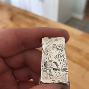 Puede incluir: Una barra de plata rectangular con una superficie rugosa y texturizada con acabado met&aacute;lico. La barra presenta hendiduras irregulares y un aspecto desgastado en su cara frontal, mostrando una est&eacute;tica cruda con brillo met&aacute;lico.
