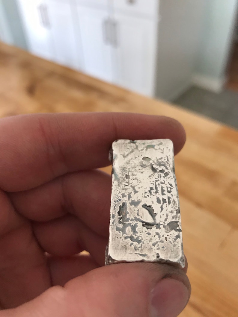 Puede incluir: Una barra de plata rectangular con una superficie rugosa y texturizada con acabado met&aacute;lico. La barra presenta hendiduras irregulares y un aspecto desgastado en su cara frontal, mostrando una est&eacute;tica cruda con brillo met&aacute;lico.