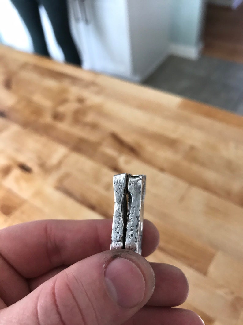 Puede incluir: Un anillo de plata con una superficie gruesa, texturizada e irregular. La banda presenta una hendidura vertical distintiva en el centro, creando una apariencia de doble capa con un acabado rugoso similar al martillado en los bordes.