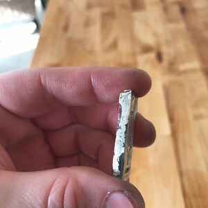 Puede incluir: Una barra de metal rectangular de color plateado con un borde rugoso y texturizado y una superficie estampada. La pieza de metal tiene un brillo met&aacute;lico y parece ser un lingote fundido con marcas visibles y lados irregulares.